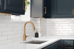 Sink detail shot in a luxury kitchen with herringbone backsplash tiles. white marble countertop, and gold faucet.